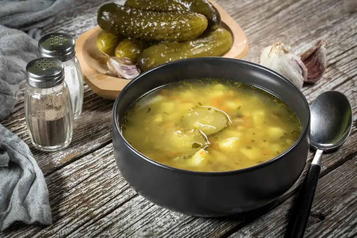 Miseczka zupy ogórkowej z ogórkami kiszonymi, czosnkiem i przyprawnikami na drewnianym stole.